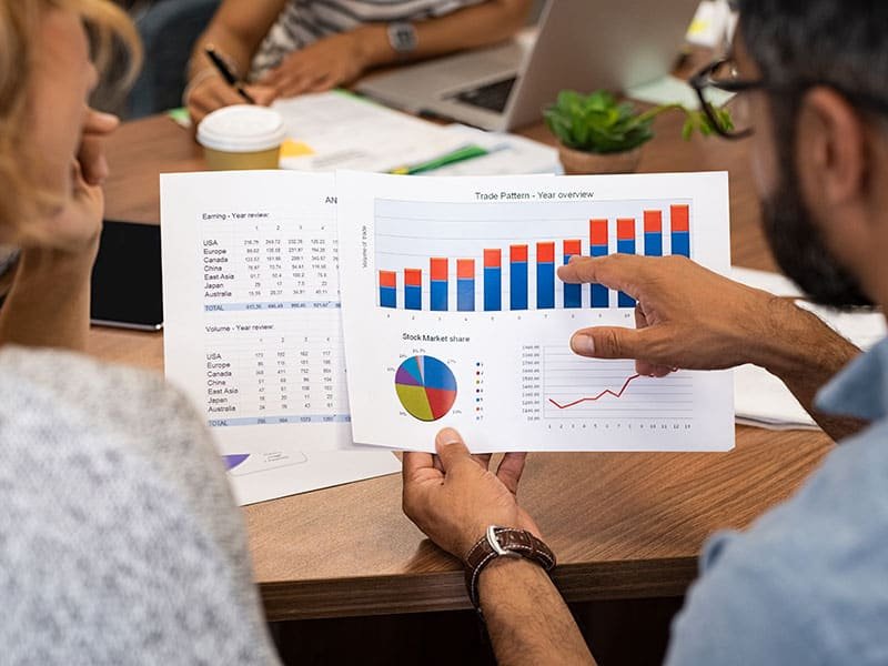 Mature latin businessman showing stock market bar graph to businesswoman. Financial advising team discussing trading patterns of year. Rear view of casual businesspeople holding charts studying year trend together.